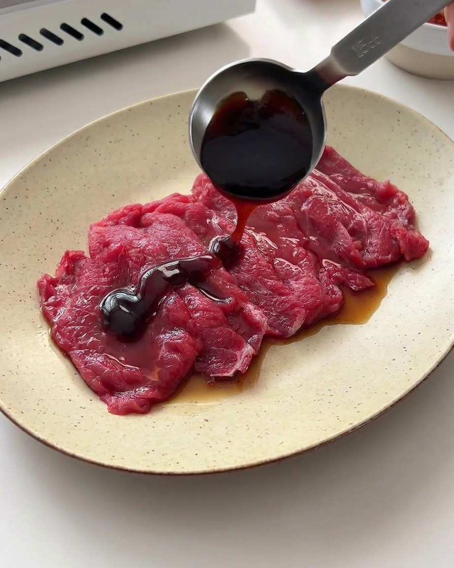 金属製の計量スプーンから、皿の上の牛肉に醤油とオイスターソースが注がれている様子。