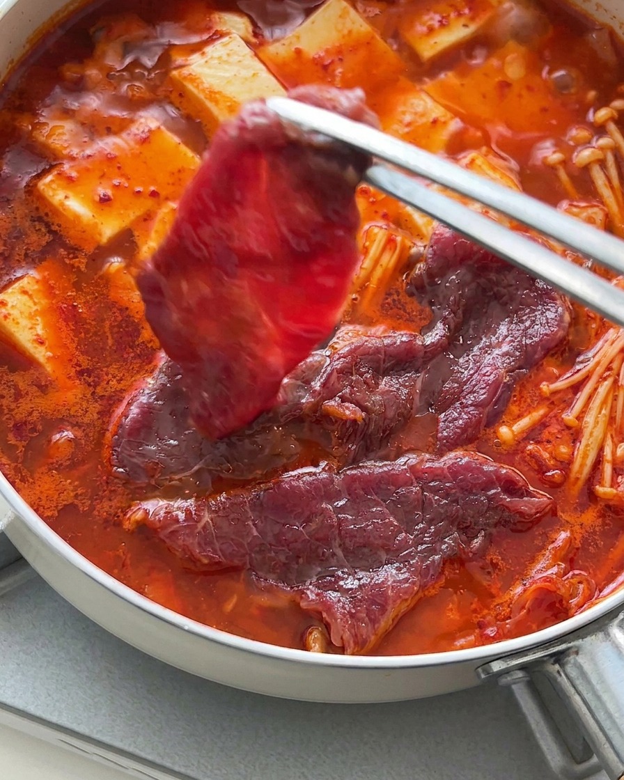 煮え立つ豆腐とキムチの上に、トングを使って生の牛肉を並べている様子。