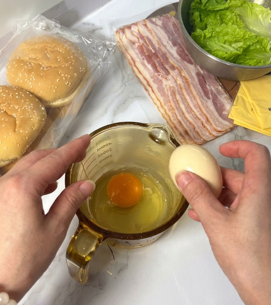 計量カップに卵を割り入れ、背後にバーガーの材料が並んでいる様子。