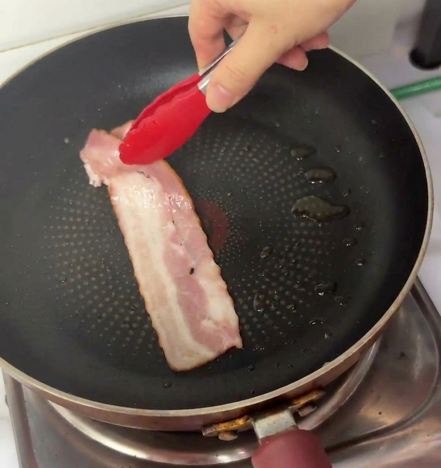 トングを使って、生のベーコンをフライパンに並べている様子。