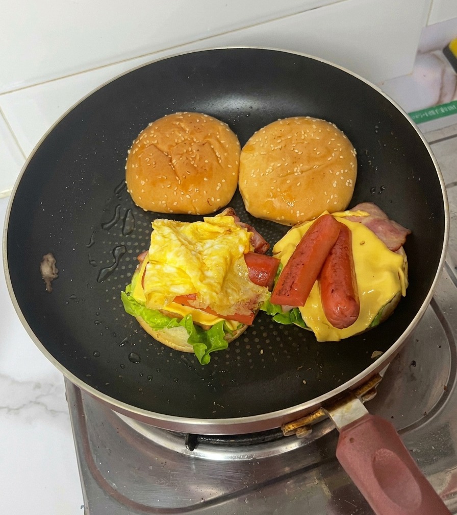 フライパンの中で、具材が積み重なったバーガーに上のバンズをのせている様子。