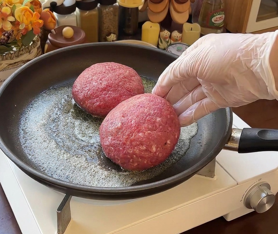 バターが溶けたフライパンに、厚みのある丸い生のビーフパティ2枚を置いている様子。右側に手袋をした手が写っている。
