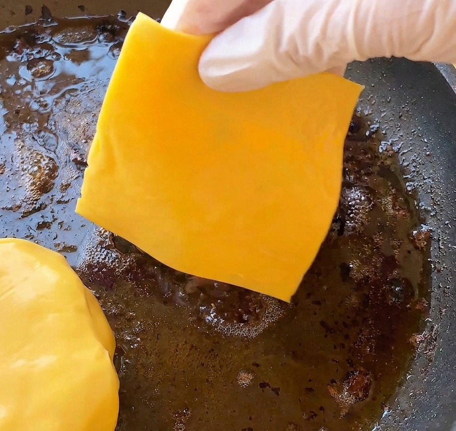 手袋をした手が、フライパンで焼かれているハンバーガーのパティの上に鮮やかな黄色のスライスチェダーチーズを乗せている様子。