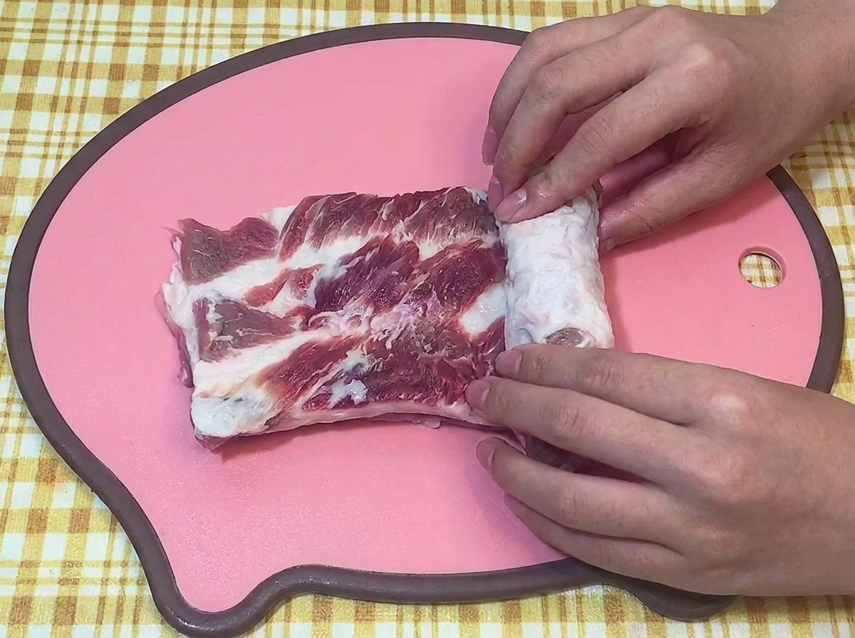 ピンクのまな板の上で、生の豚バラ肉を両手でしっかりと円柱状に巻いている様子。