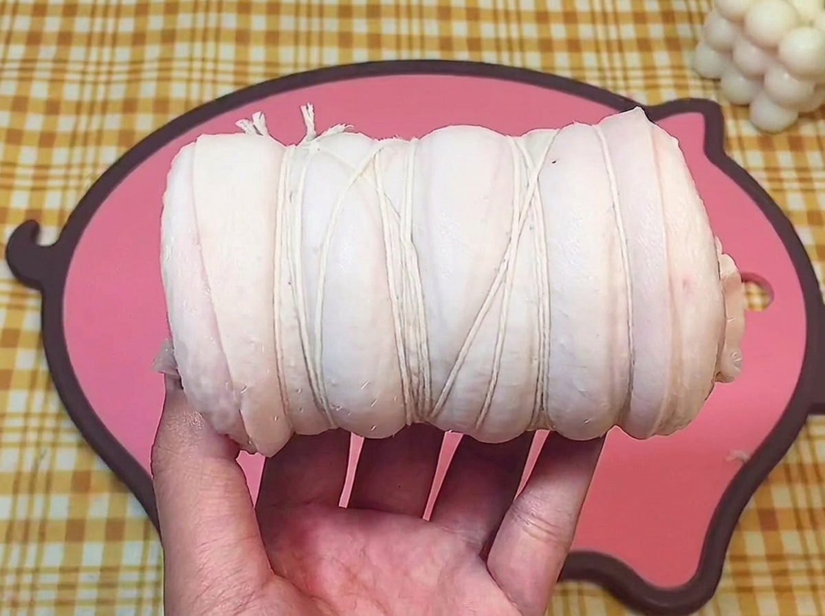 ピンクのまな板の上で、丸めた豚バラ肉を白いタコ糸で等間隔に縛っている様子。