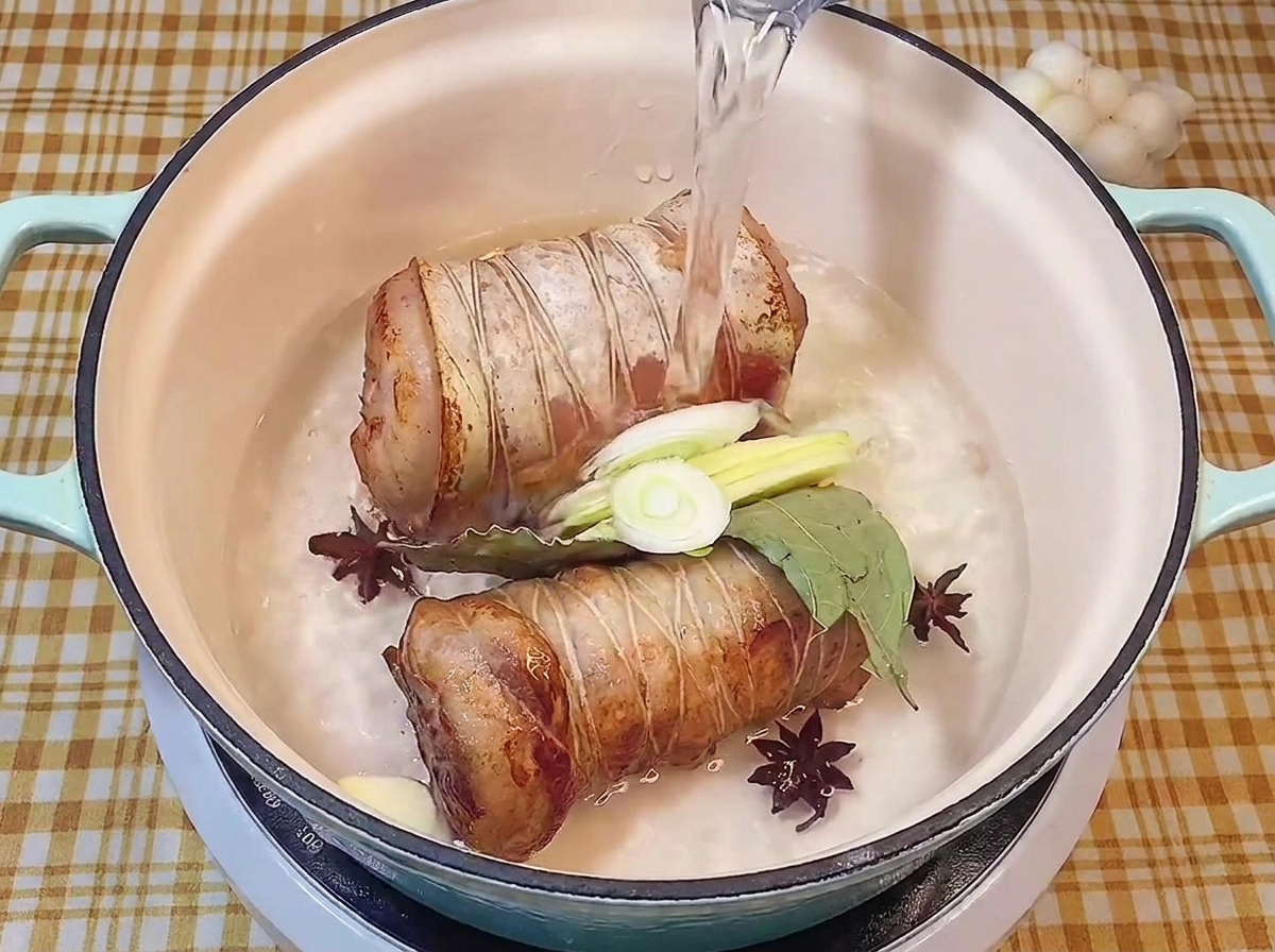 鍋に入れた焼き色のついた豚肉に、ネギ、生姜、にんにく、八角、ローリエを加え、水を注いでいる様子。
