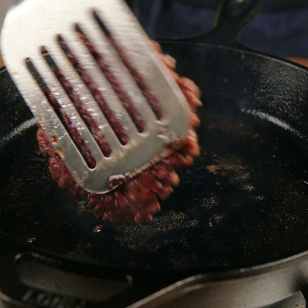 金属製のヘラで、表面がカリカリに焼けた肉をスキレットの中で一気にひっくり返している様子
