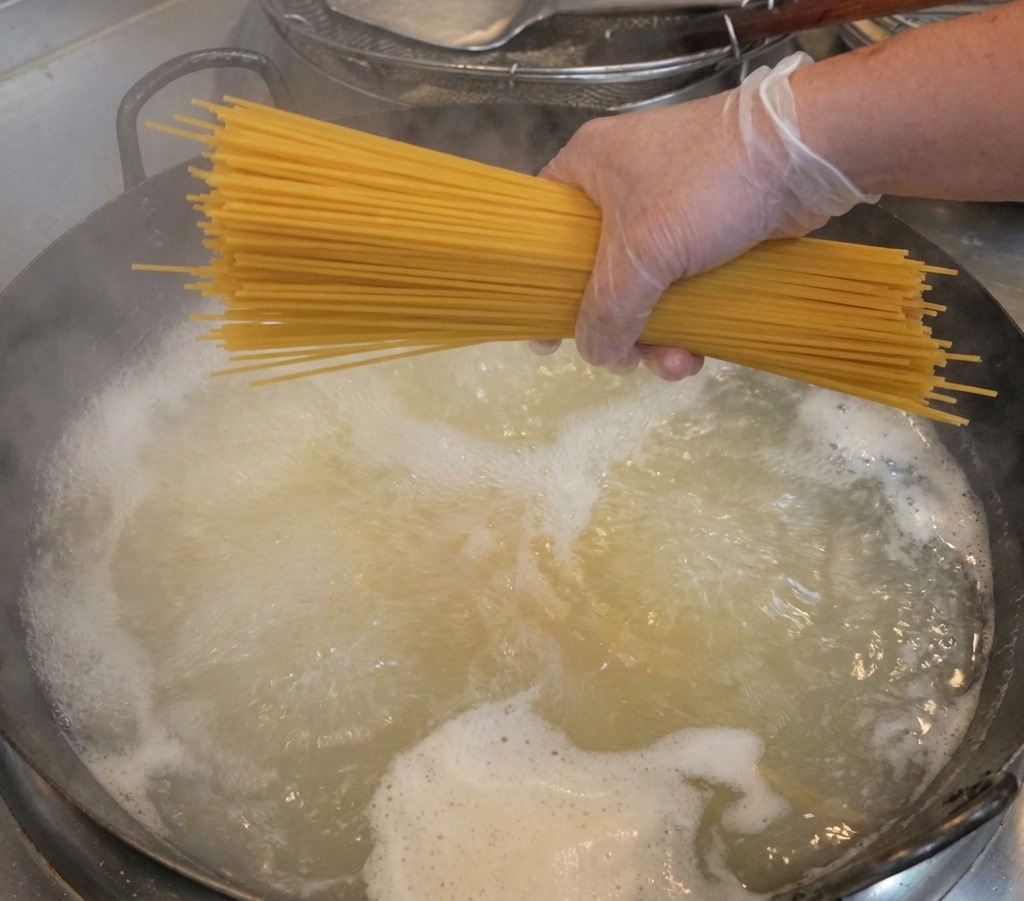 手袋をした手で、乾燥したスパゲッティの束を沸騰した鍋の上にかざしている様子。
