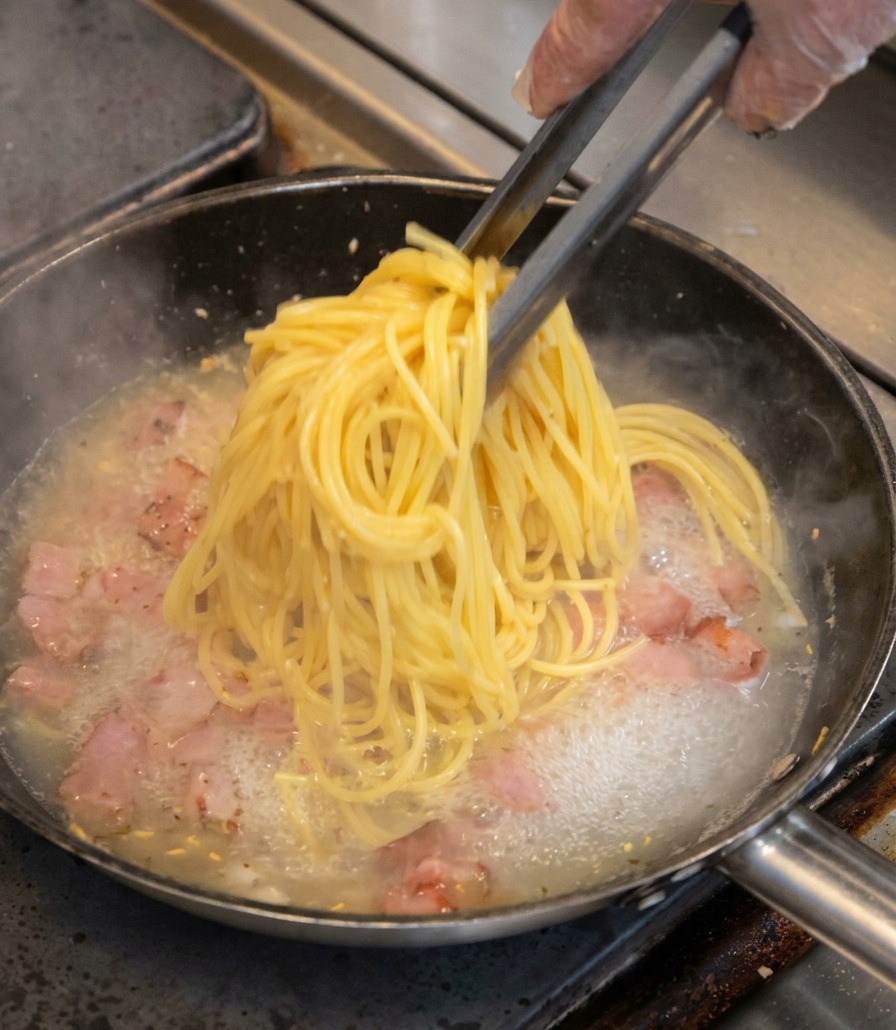 トングで掴んだスパゲッティを、ベーコンの入ったフライパンに移している様子。