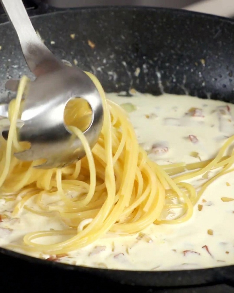 茹で上がったスパゲッティをトングでクリームソースの入ったパンに移す様子