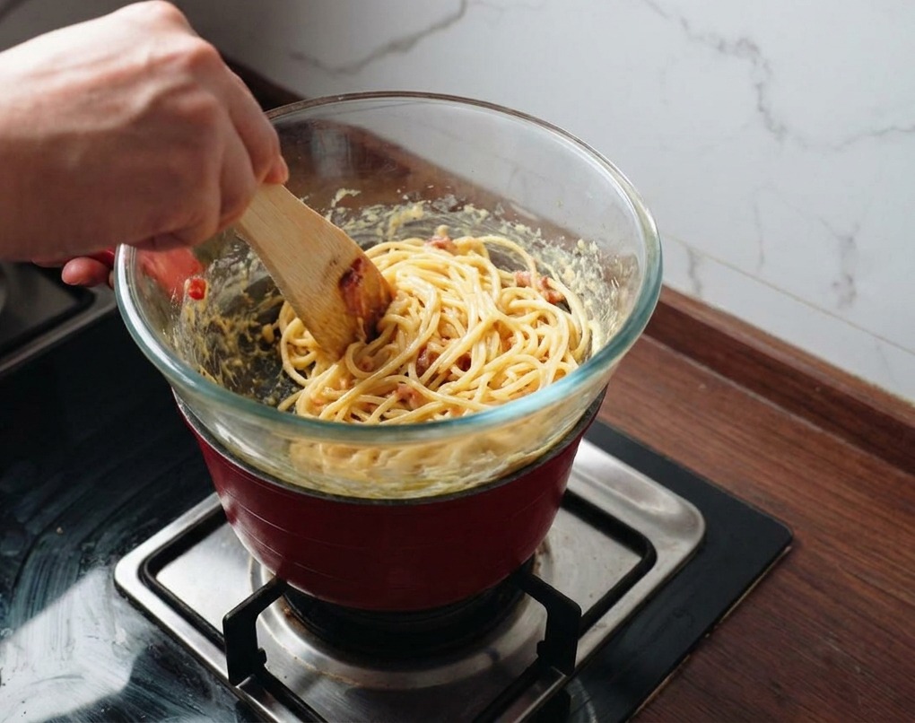 湯気が上がる鍋の上で、スパチュラを使ってパスタとソースを激しく混ぜ合わせている様子。