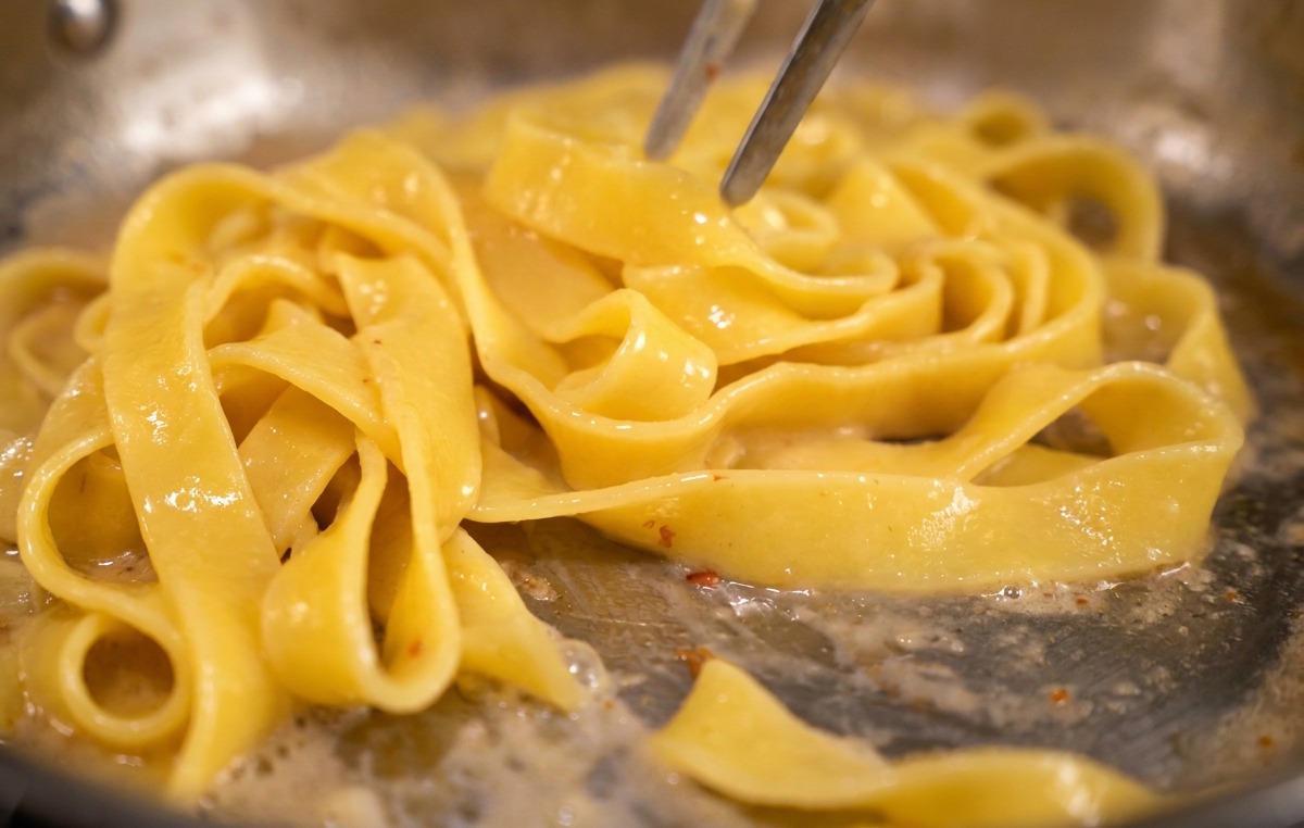 トングでパスタを混ぜながら、黄色い卵ソースを回し入れている様子。