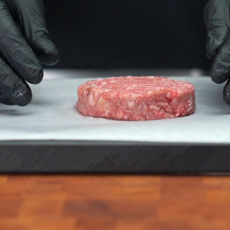 白いクッキングシートの上に置かれた生の牛肉パティ。表面に黒こしょうの粒が見える。