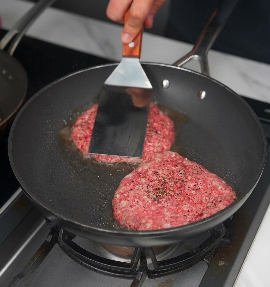 金属製のヘラを使って、フライパンの中の2枚のパティを強く押し付けて焼いている様子。