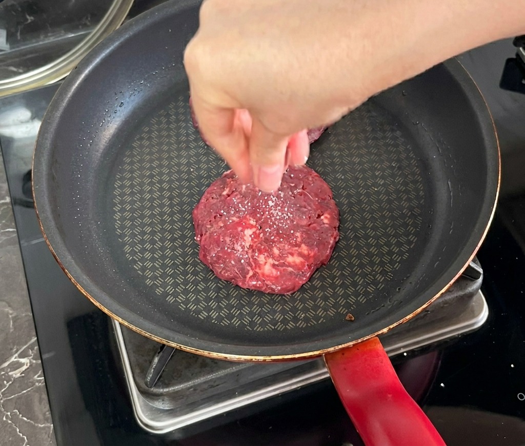 フライパンで焼いているパティの上から、高い位置で塩を振っている様子