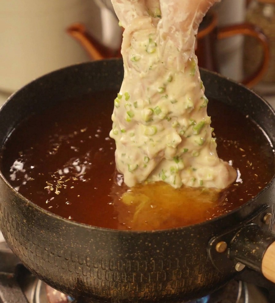 透明なビニール手袋をした手で、万能ねぎの衣をたっぷり纏った鶏肉を熱い油が入った深い鍋にそっと入れている様子。