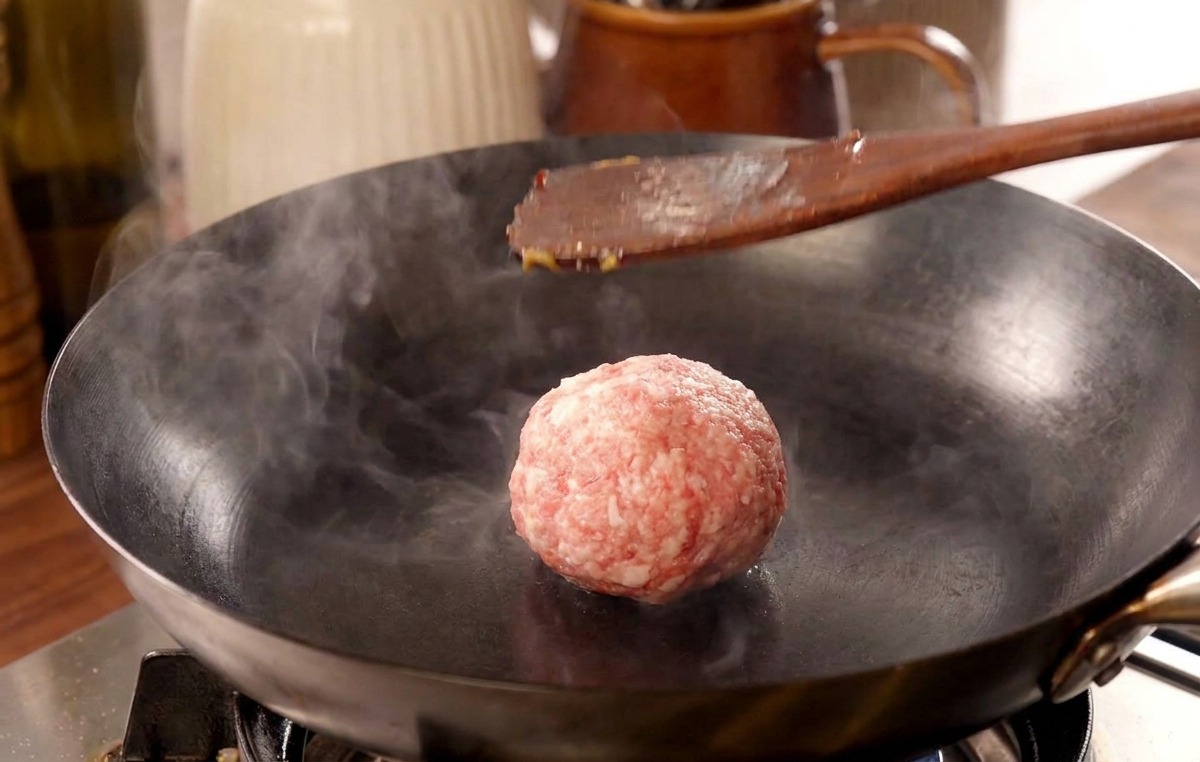 熱いフライパンの上で、ヘラを使って肉のボールを強く押し潰している様子。