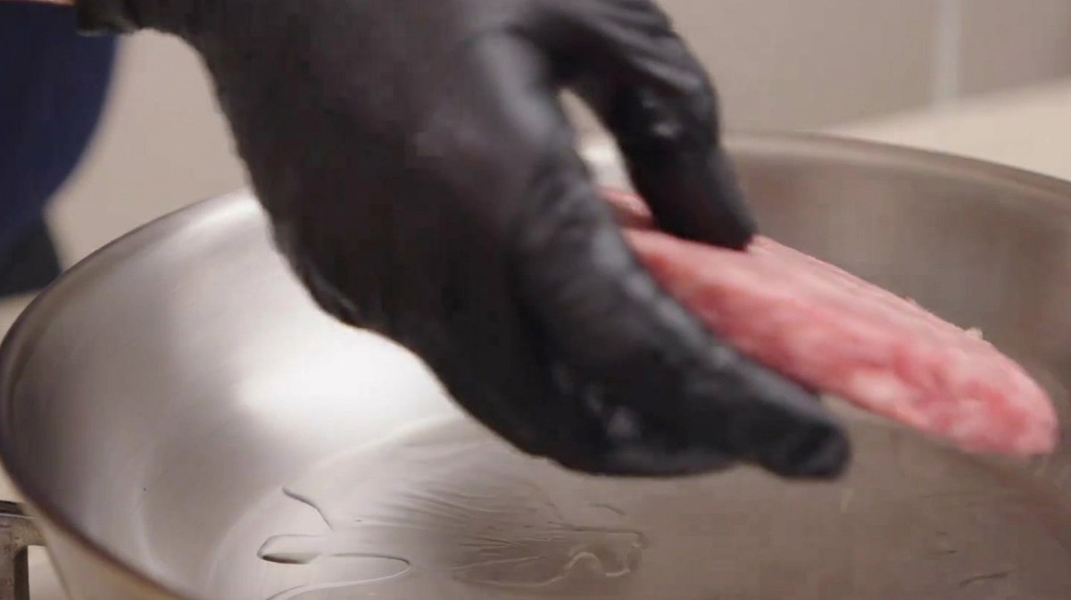 黒い手袋をはめた手が、油を引いたステンレススキレットにサシの入った生の和牛パティを丁寧に入れている様子。
