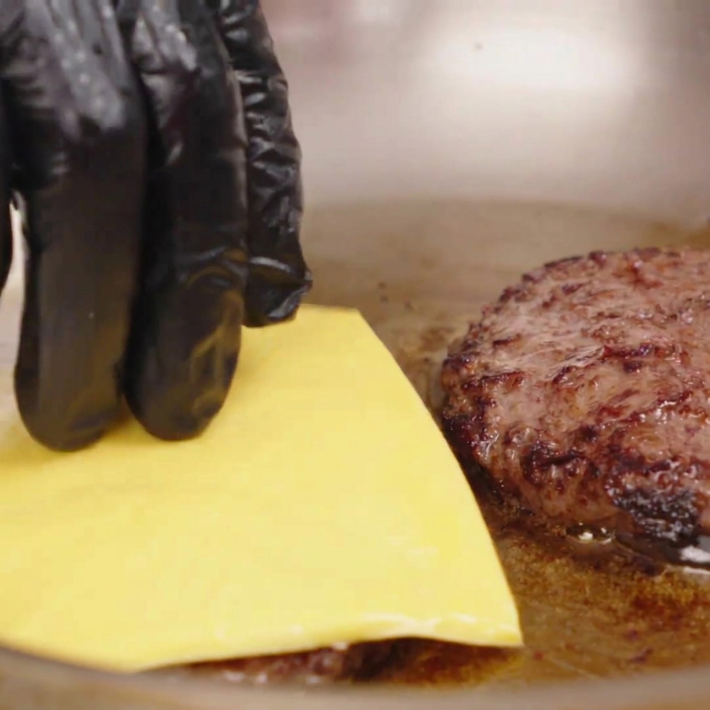 フライパンの中の焼きたての肉パティの上に、四角い黄色いチーズを置いている様子。
