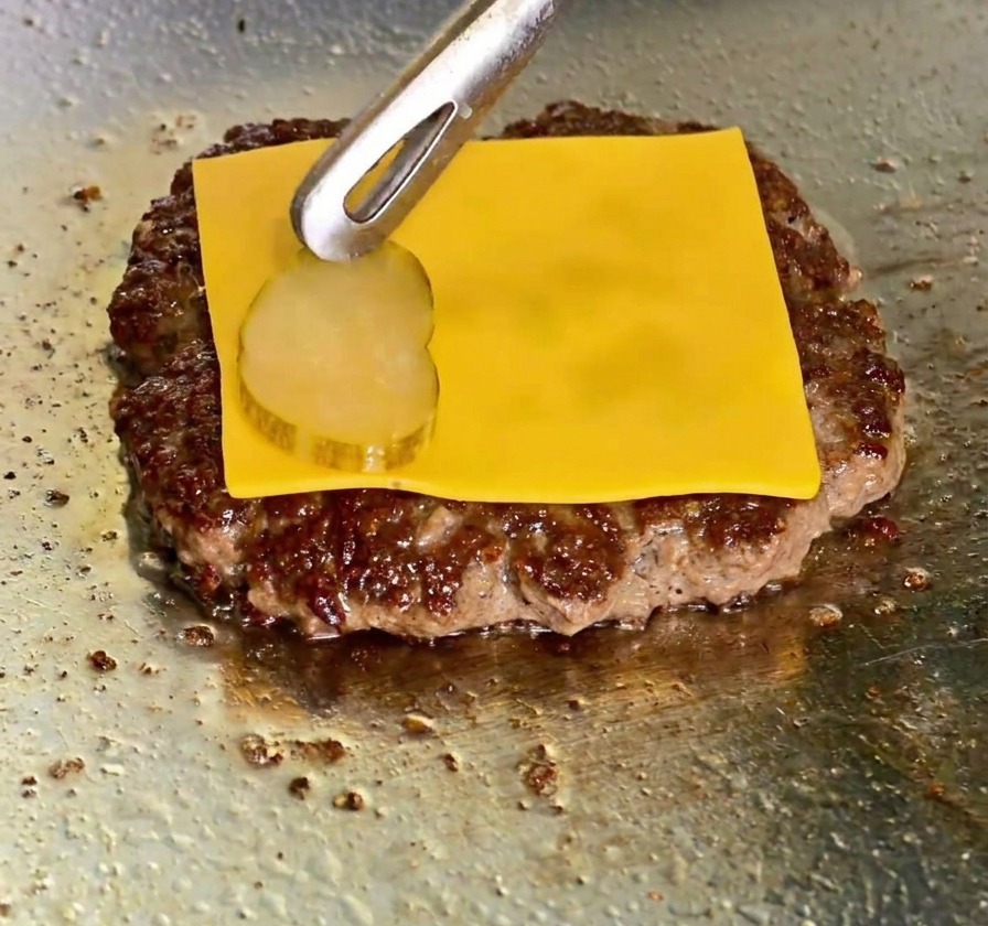 パティの上にのせられたチェダーチーズが溶け始め、トングでその上にピクルスが置かれている様子。