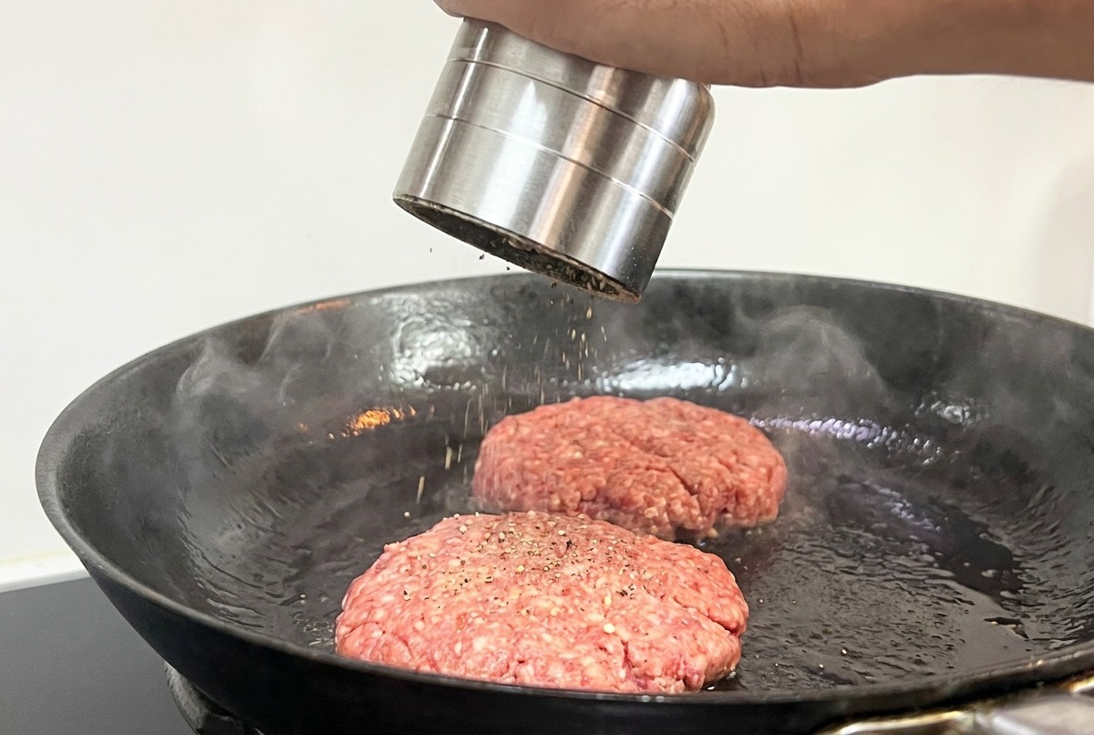 フライパンで焼いている肉に、ペッパーミルで黒胡椒を振りかけている様子。