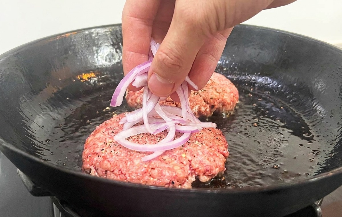 フライパンの中の生肉の上に、スライスした玉ねぎを乗せている様子。