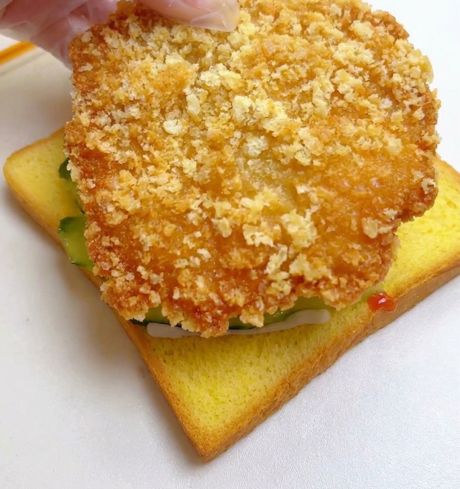 きゅうりをのせたパンの上に、大きく黄金色に揚がったチキンフィレをのせている手。