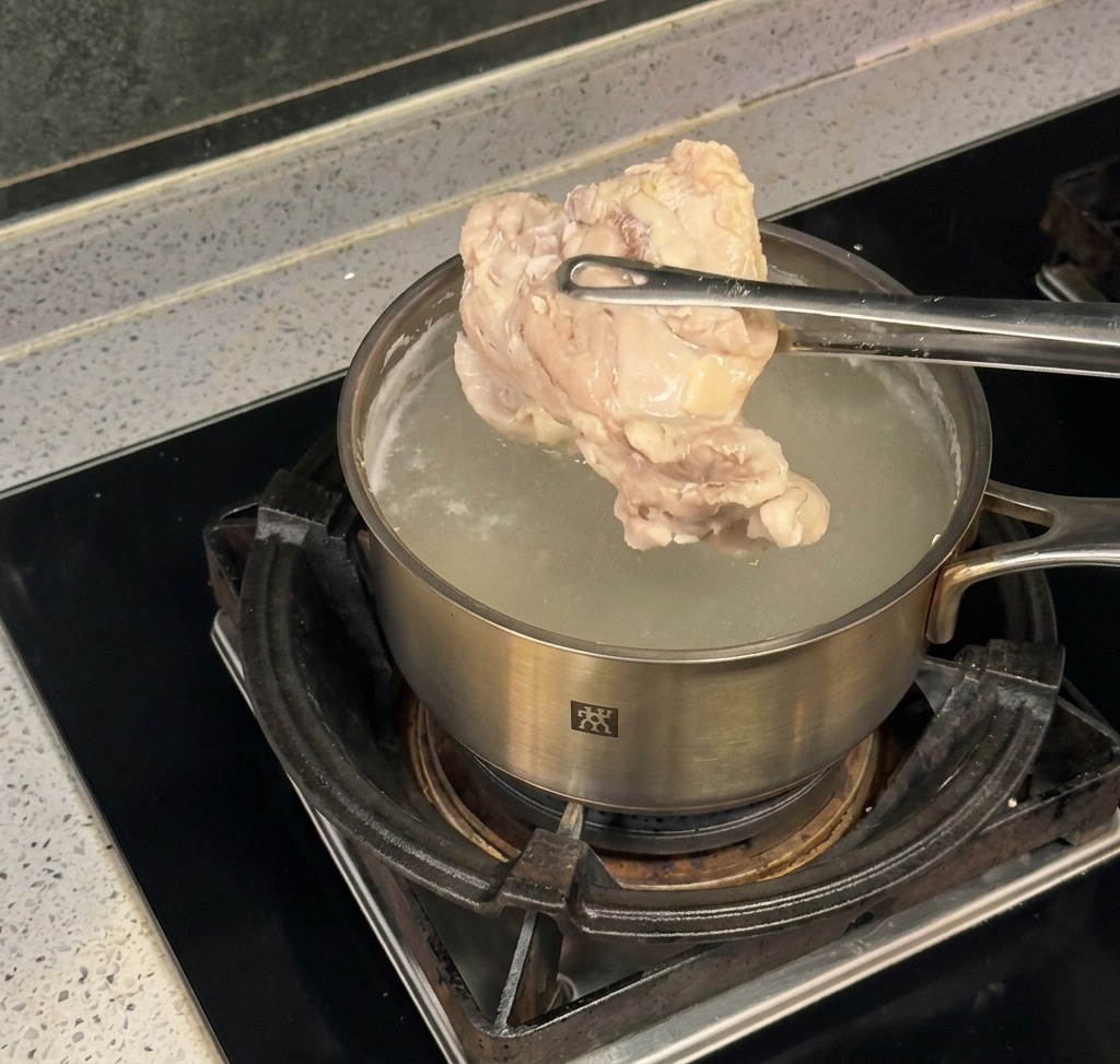 コンロの上の鍋で茹で上がった大きな鶏肉を、トングで持ち上げている様子。