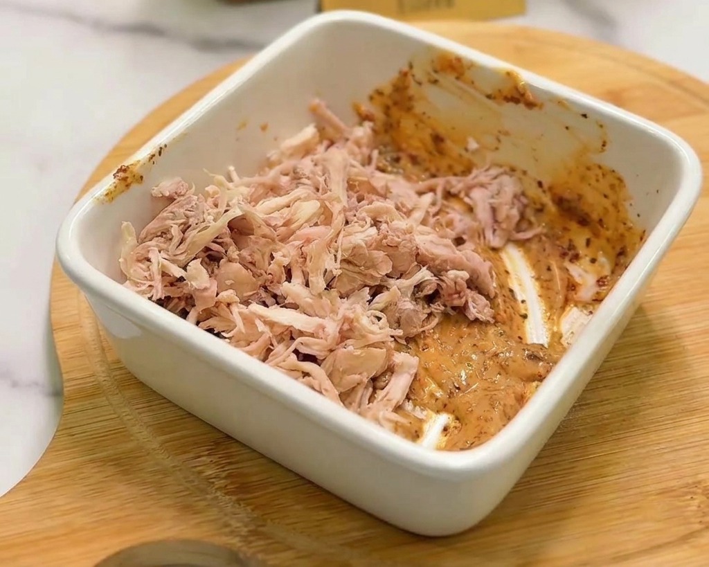 ほぐした鶏肉を、スパイシーなオレンジ色のソースが入ったボウルに入れている様子。