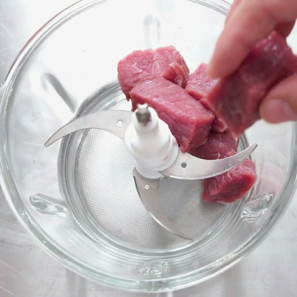 生の牛肉の塊を、マルチブレードが付いたガラス製のフードチョッパーに入れている様子。