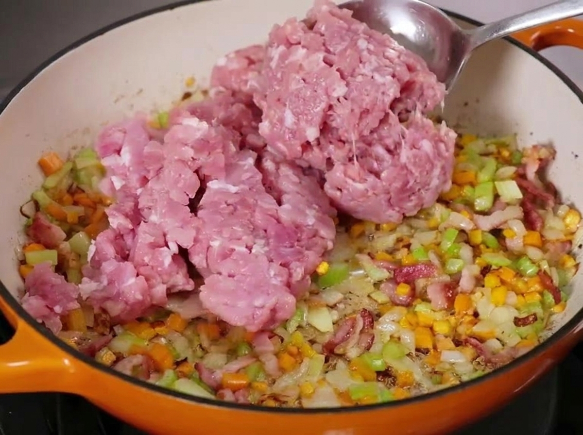 野菜とベーコンを炒めたフライパンに、生のひき肉を大きなスプーンで加えている様子。