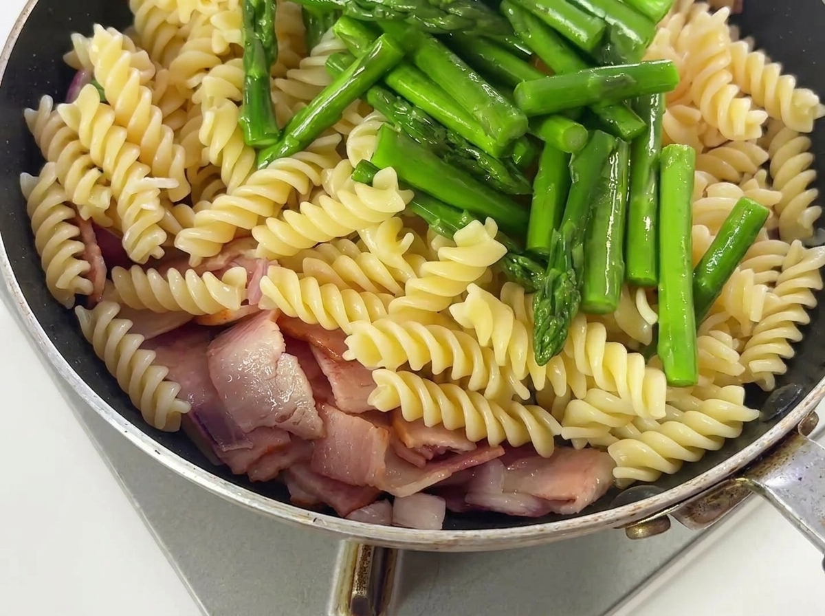 フライパンの中でアスパラガスと調味料をパスタと和えている様子。
