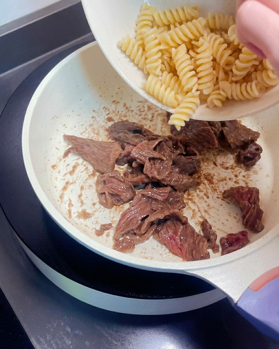 茹でたスパイラルパスタを、炒めた牛肉の入ったフライパンに入れている様子。