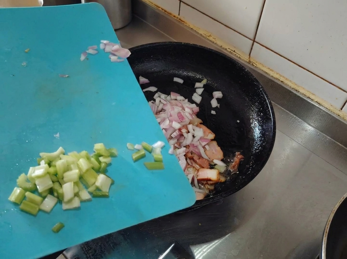 みじん切りにしたエシャロットとセロリを、焼き色のついたベーコンの入ったフライパンに加える様子。