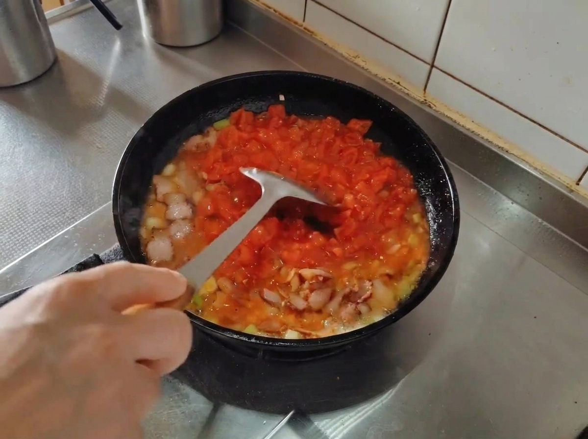 フライパンの中でベーコンや野菜と一緒に煮込まれているカットトマトを、金属製のヘラでつぶす様子。