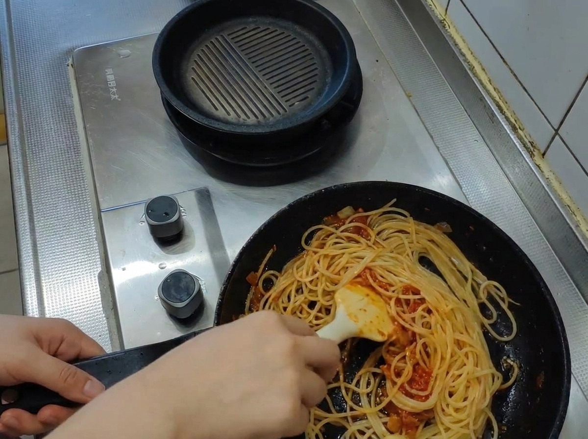 お玉で白濁した熱い茹で汁を、スパゲッティとトマトソースの入ったフライパンに注ぐ様子。