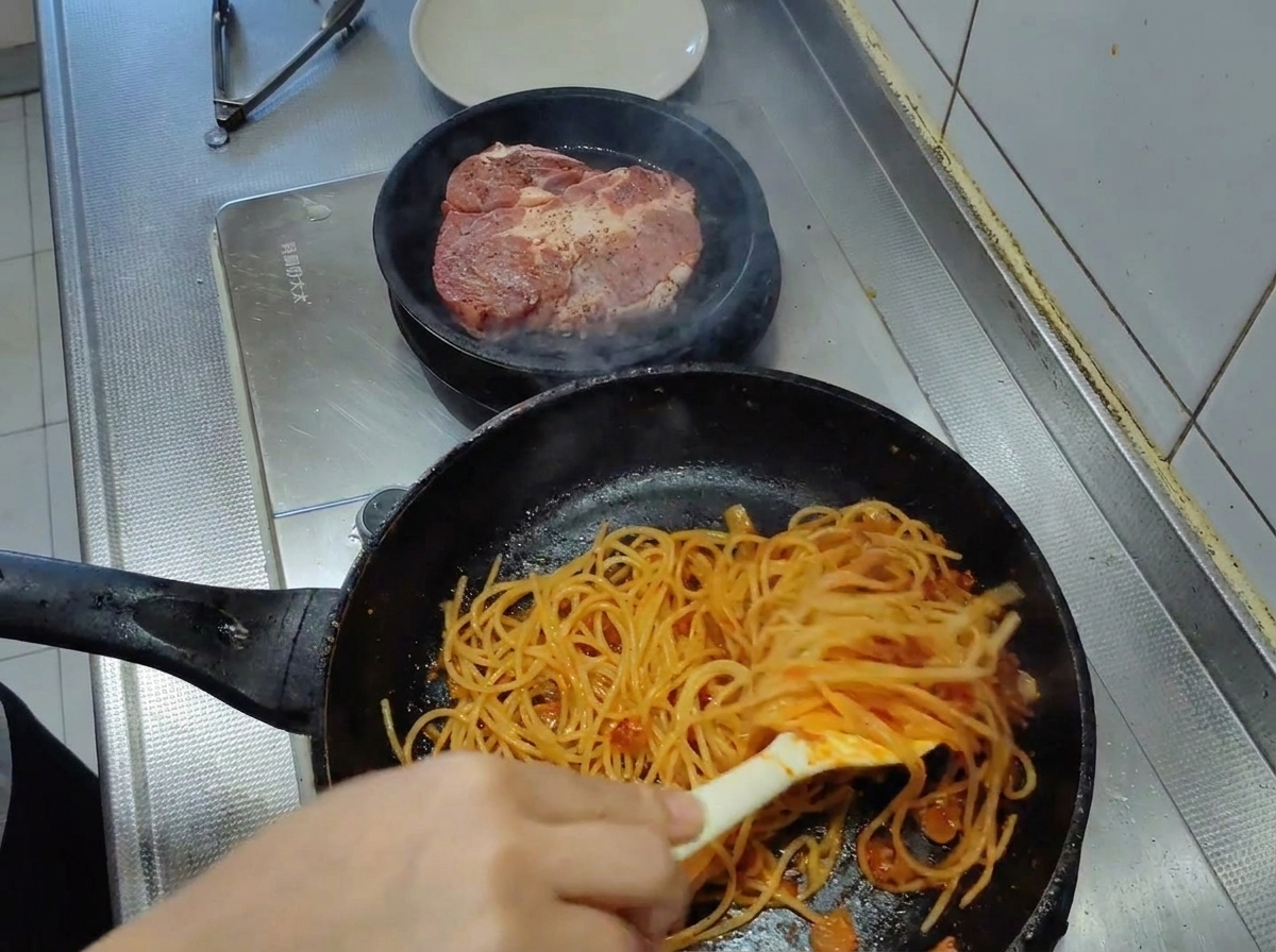 フライパンの中でスパゲッティをトングで混ぜ合わせ、濃厚で色鮮やかなトマトソースを全体に絡める様子。