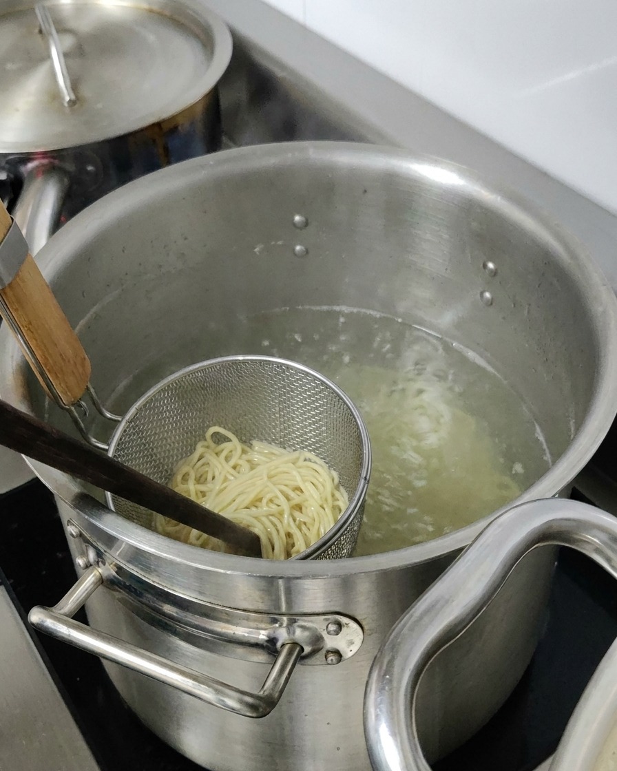 大きな鍋の中で、金属製のザルに入ったラーメンの麺を茹でている様子。