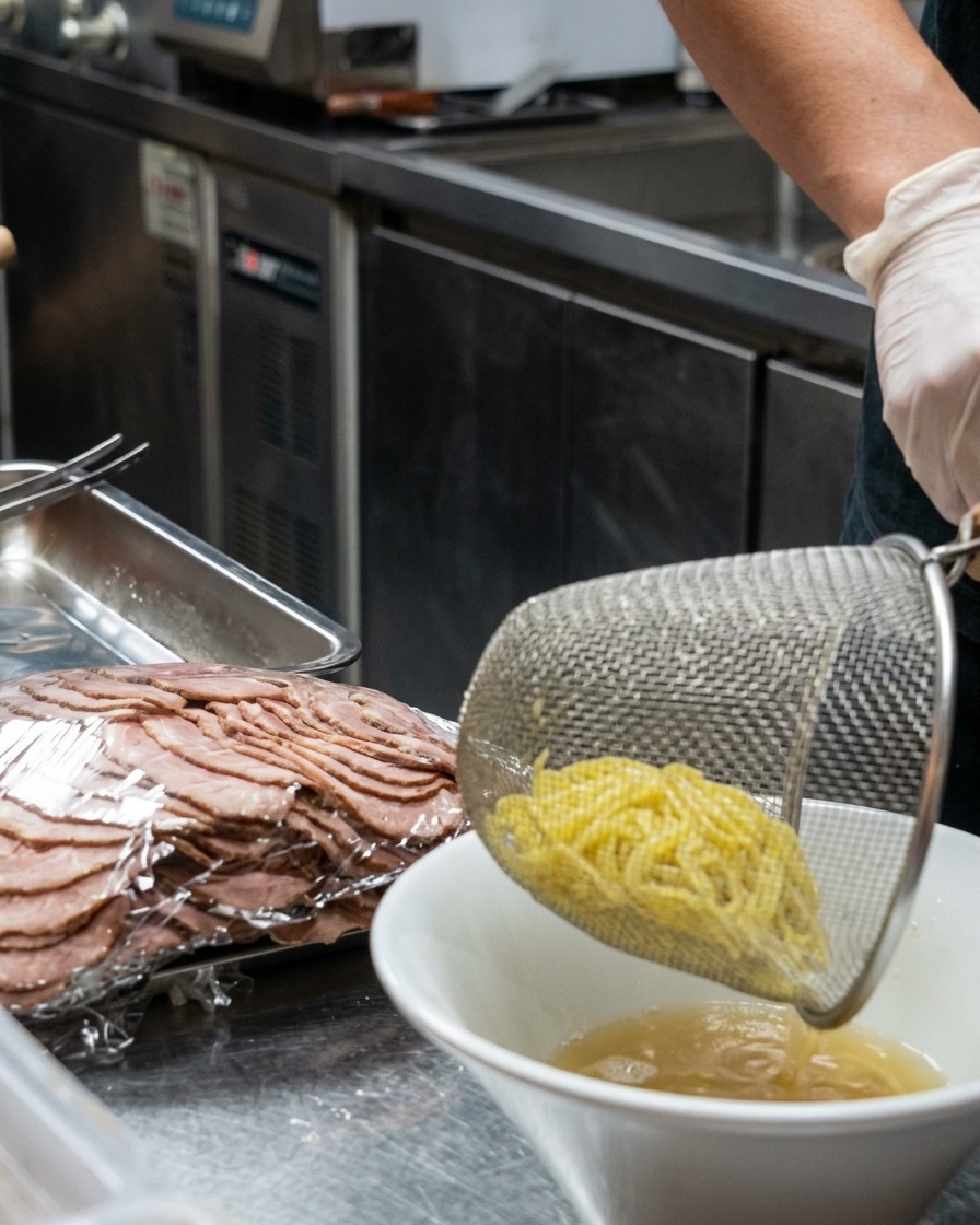 茹で上がったばかりの黄色いラーメンの麺を、湯切りザルからクリーミーなスープの入った白い器に移している様子。