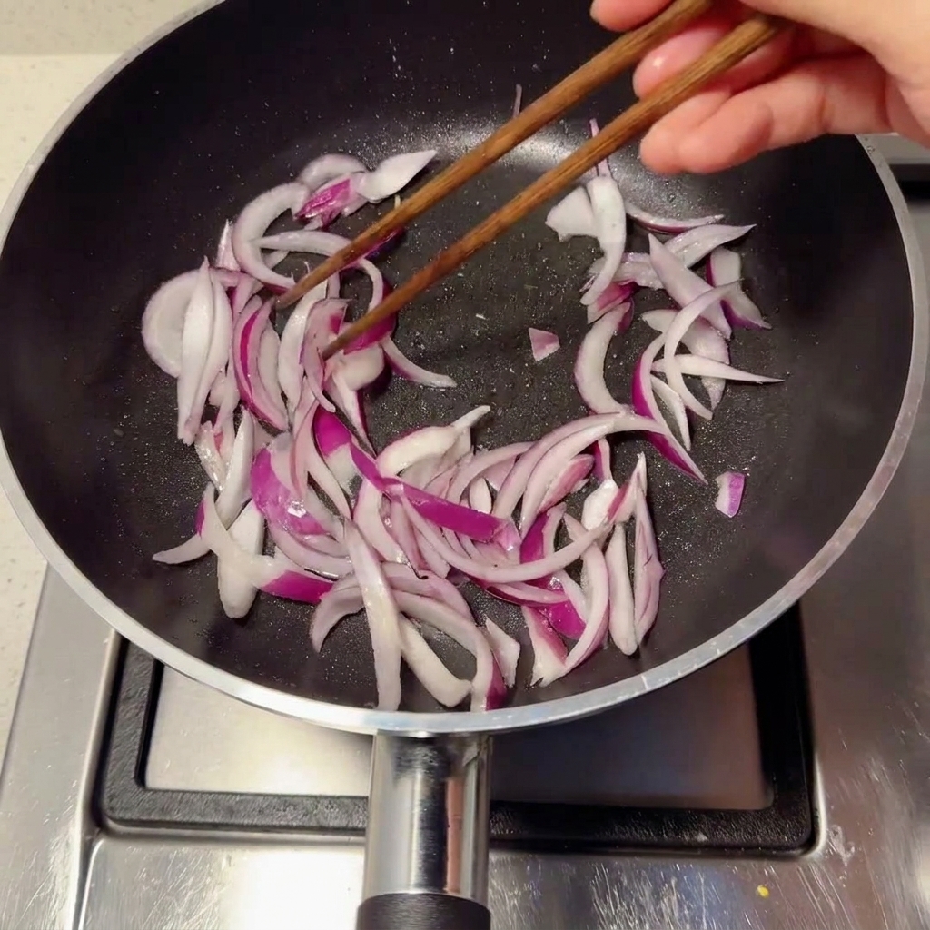 黒いフライパンでスライスした紫玉ねぎを菜箸で炒めている様子。