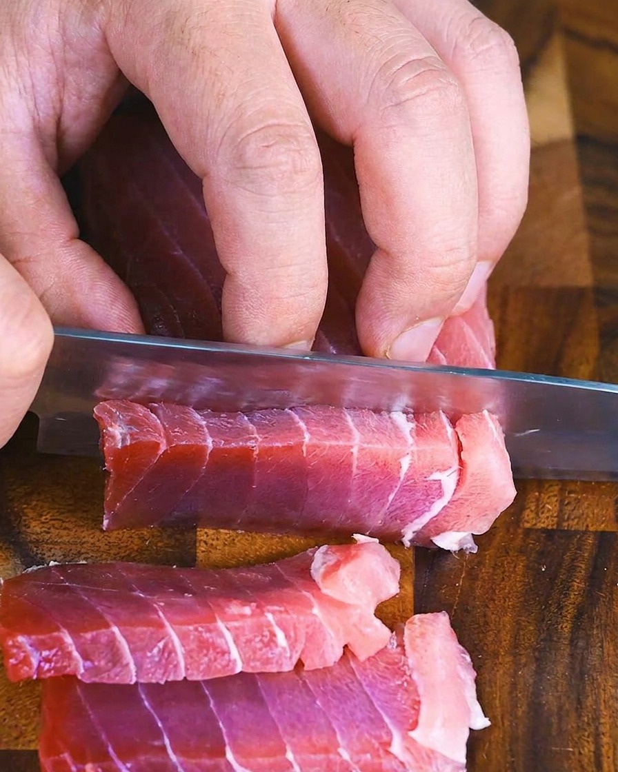 木のまな板の上で、包丁を使って赤い生のマグロの柵を厚めの平切りにしている手元の様子。