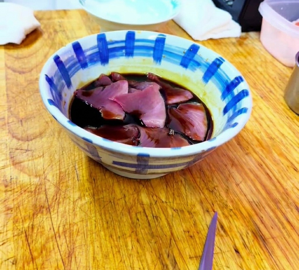 陶器のボウルの中で、マグロの切り身が醤油ベースの漬けダレに浸っている様子。