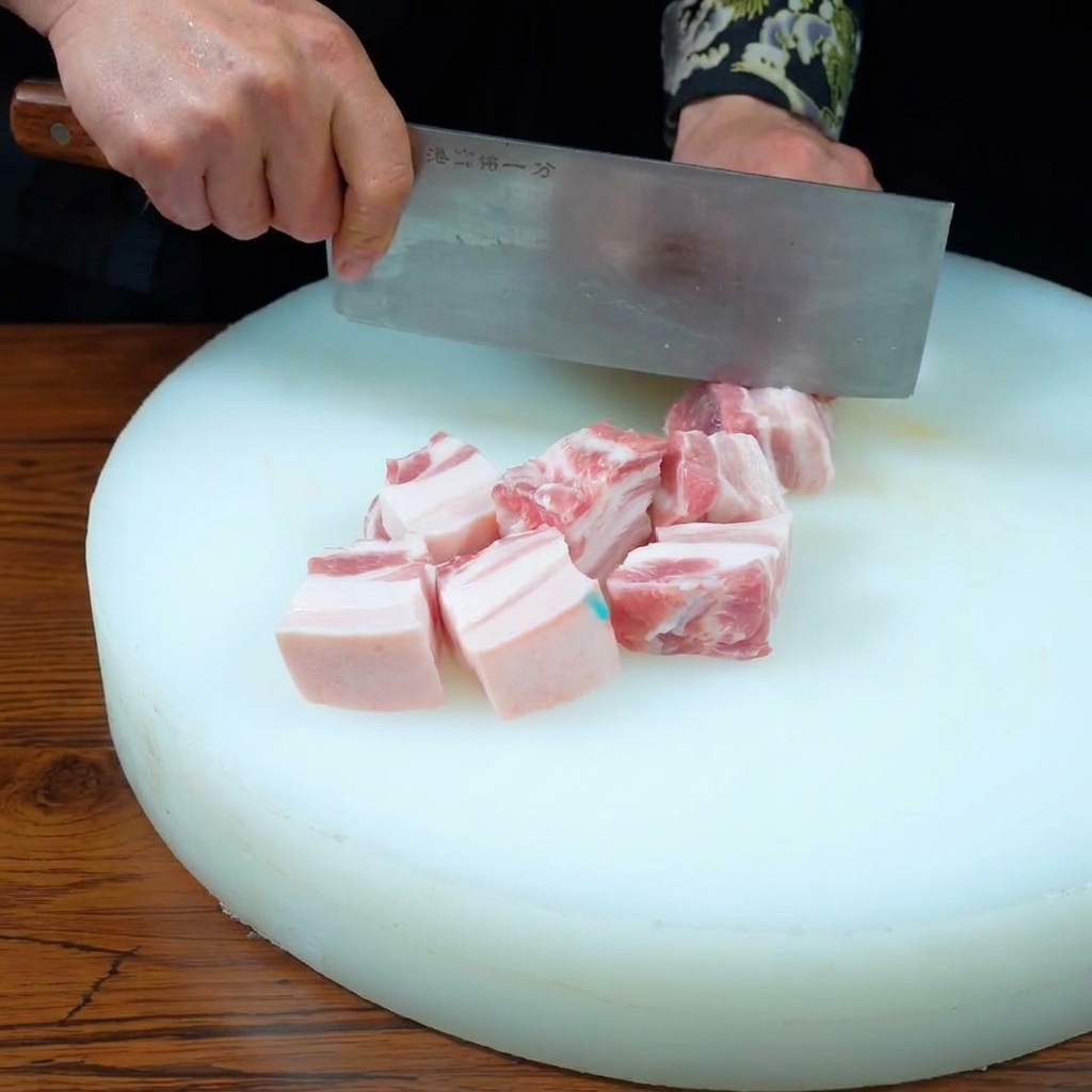 白いまな板の上で、包丁を使って三枚肉を均等なサイコロ状に切る様子。