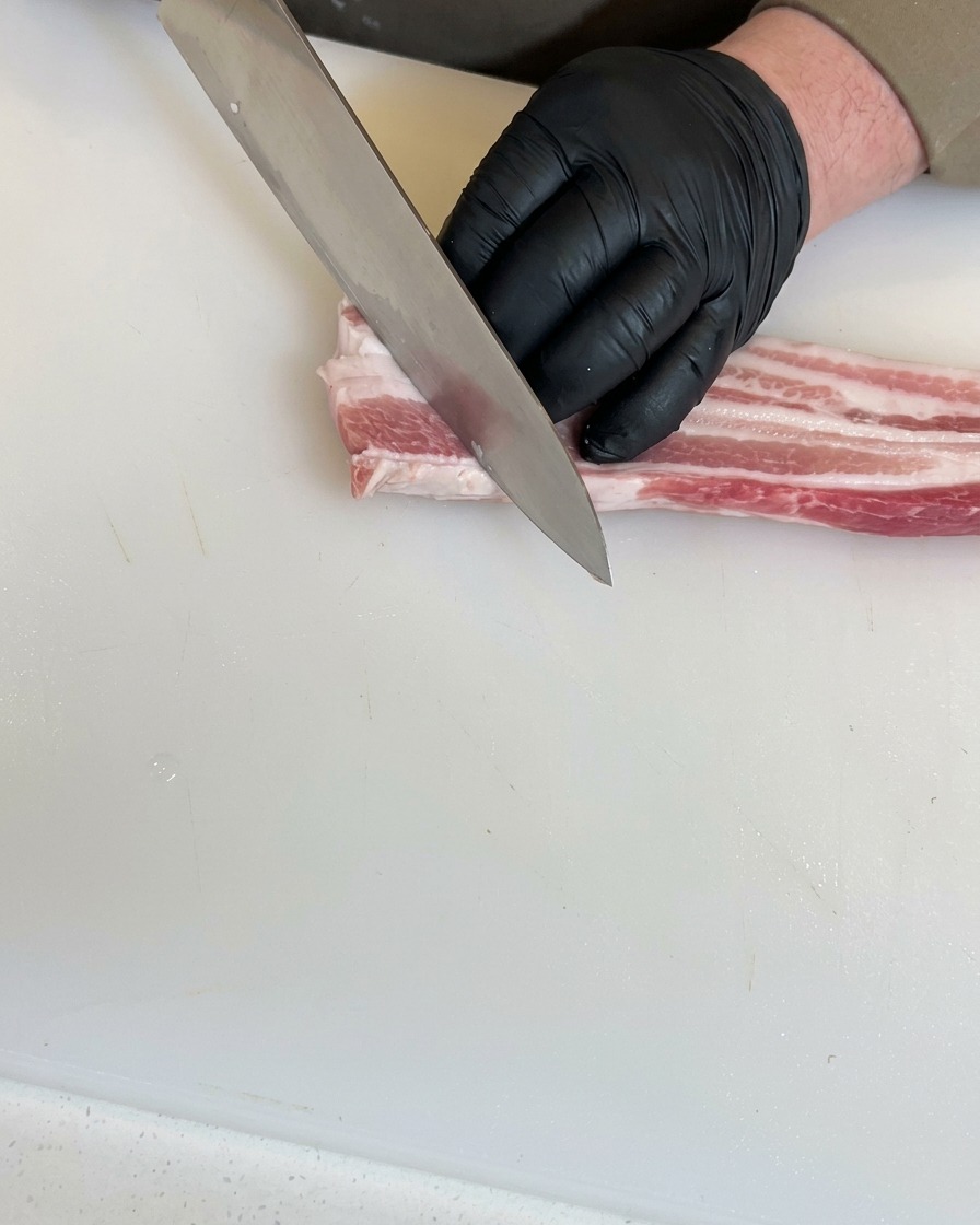 黒い手袋をした手が、まな板の上の生の豚バラ肉にシェフナイフで斜めに深い切り込みを入れている様子。