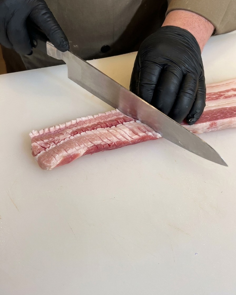 黒い手袋をした手が、豚バラ肉の裏面に格子状の深い切り込みを入れている様子。