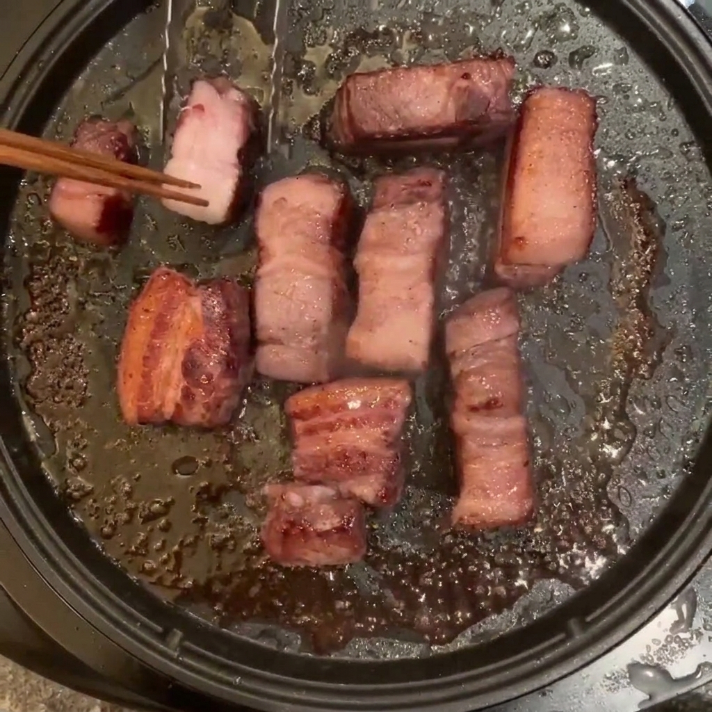 ホットプレートで焼かれている豚バラ肉を、木の箸で裏返している様子。