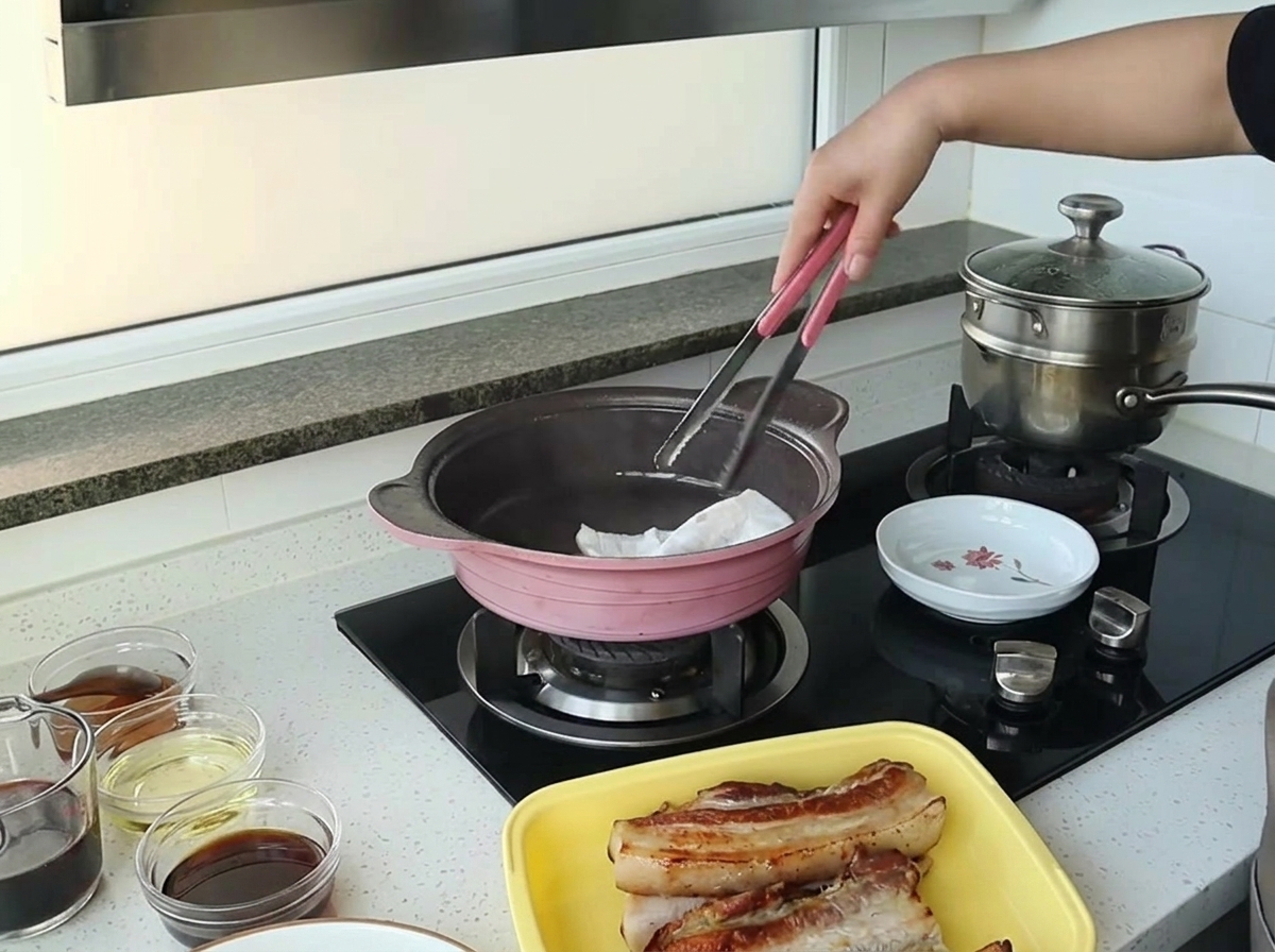 ピンクのフライパンの余分な油を、折りたたんだキッチンペーパーをトングで挟んで拭き取っている様子