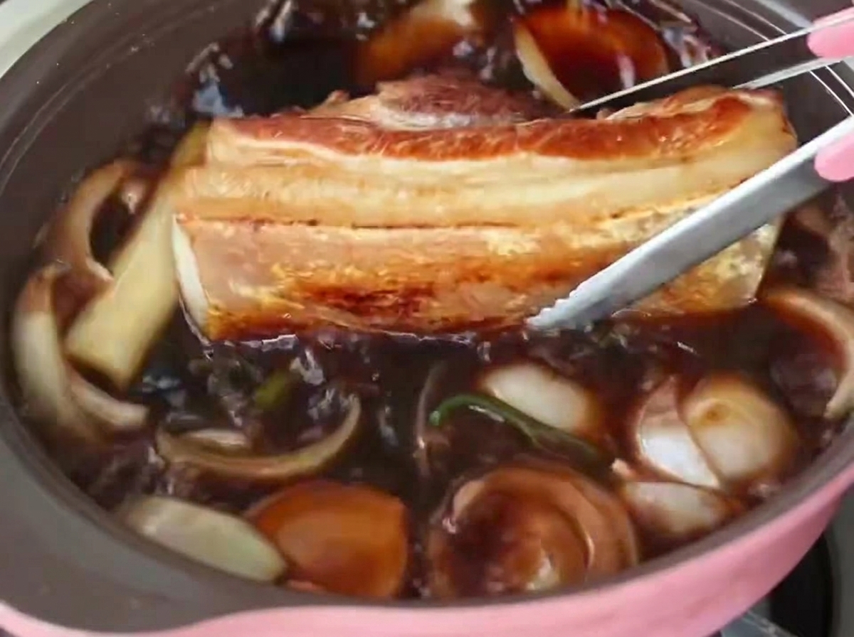 焼いた豚バラ肉の厚切りを、玉ねぎが入った煮汁の沸立つ鍋にトングで戻し入れている様子