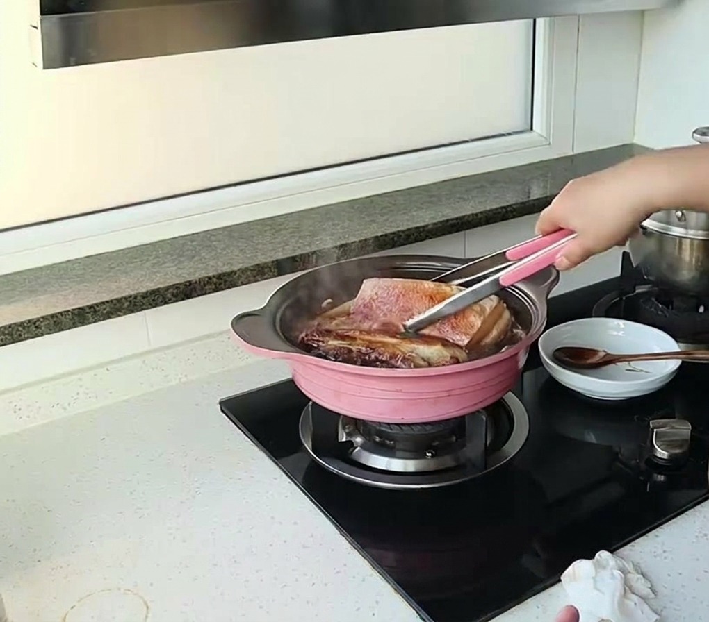 ピンクの鍋に入った濃い色のソースの中で煮込まれている大きな豚バラ肉を、トングを使って裏返している様子
