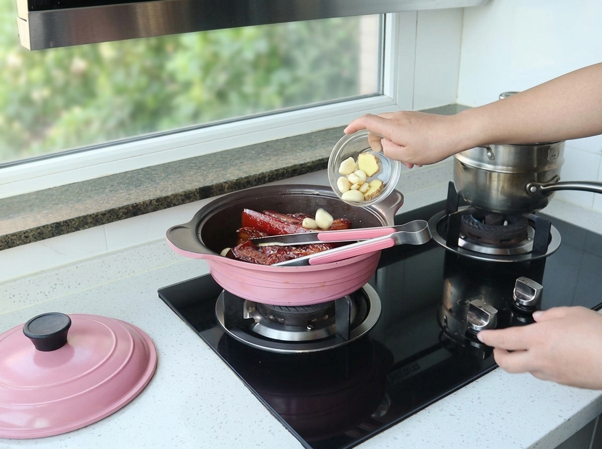 煮込まれた豚バラ肉が入ったピンクの鍋に、小さなガラスの器から皮をむいたにんにくと生姜のスライスを入れている様子